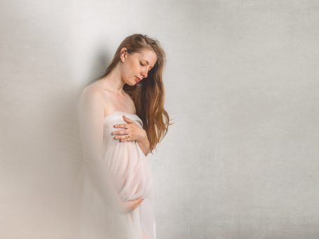 Mujer embarazada con vestido claro, apoyada en una pared, mirada suave y contemplativa.