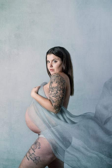 Mujer embarazada con tatuajes, sosteniendo una tela que acaricia su abdomen.