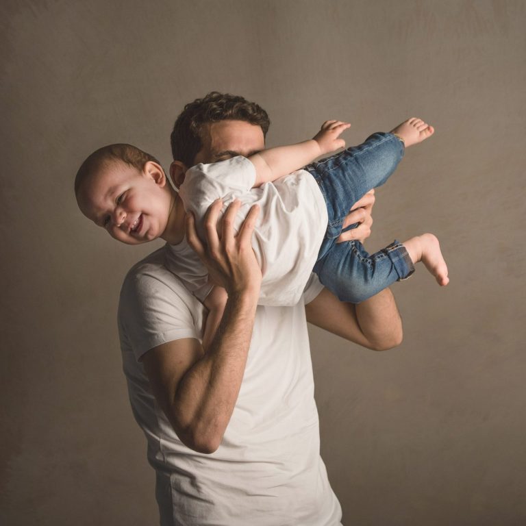 Hombre sostiene a un niño pequeño, ambos sonrientes, contra un fondo claro.