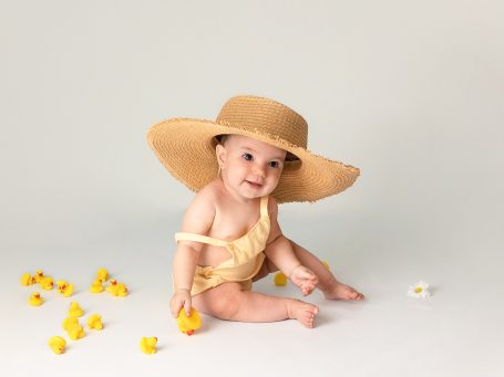 Bebé sonriente con sombrero grande, sentado entre flores amarillas.