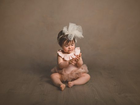 Niña pequeña sentada con un vestido rosa y un lazo en la cabeza, sosteniendo un dulce.