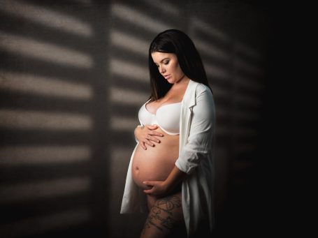 Mujer embarazada en lencería, acariciando su abdomen bajo luz suave y sombras.