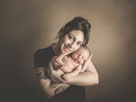 Mujer sonriente sostiene a un bebé desnudo en sus brazos, en un fondo neutro.