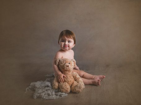 Niño pequeño sentado en el suelo, abrazando un oso de peluche. Fondo neutro.