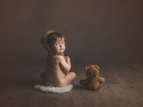 Niña pequeña mirando hacia atrás, sentada sobre una alfombra, con un osito de peluche al lado.