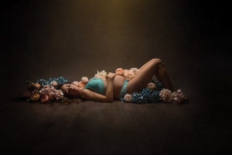 Mujer embarazada reclinada entre flores, con cabello azul y bañador turquesa.