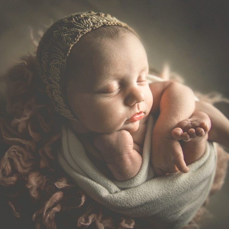 Bebé dormido con gorro y piel clara, rodeado de mantas suaves y rosas.