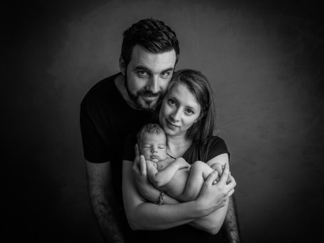 Pareja sosteniendo a un bebé en brazos, todos sonriendo en un fondo oscuro.