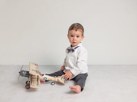 Niño pequeño sentado en el suelo con una juguete de avión, viste camisa blanca y pajarita.