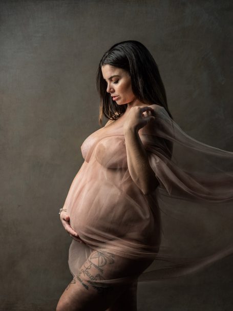 Mujer embarazada con tela transparente, posando de perfil en un fondo neutro.