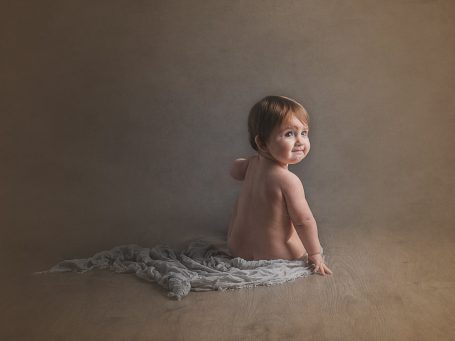 Un bebé desnudo sonríe mientras está sentado sobre un fondo marrón claro.