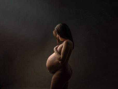 Mujer embarazada de perfil, en un ambiente oscuro, con luz suave resaltando su figura.