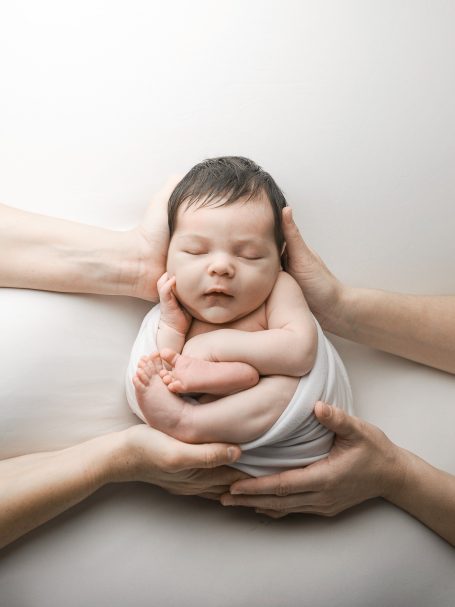Bebé dormido envuelto en una manta, sostenido con manos de adultos.