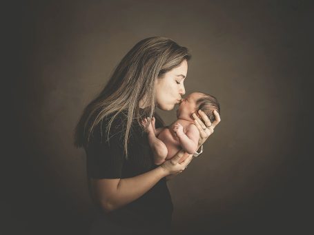 Mujer besando a un bebé en brazos, fondo oscuro y acogedor.