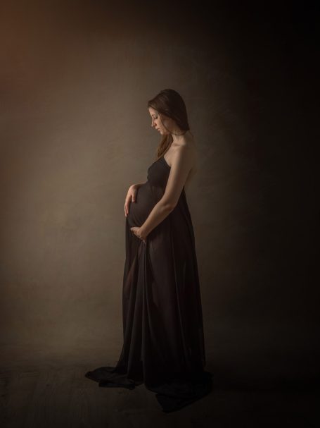 Mujer embarazada de pie, sosteniendo su vientre, con una vestido negro en un fondo suave.