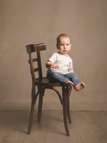 Niño pequeño sentado en una silla de madera, con una expresión curiosa y pies descalzos.