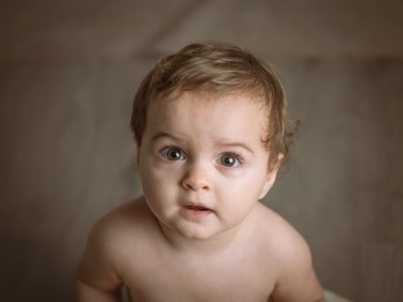 Bebé de cabello rizado, con mirada curiosa, sentado sobre una superficie clara.