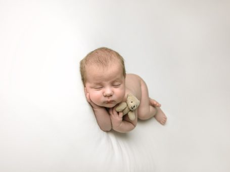 Bebé dormido en posición fetal sosteniendo un peluche, sobre fondo blanco.