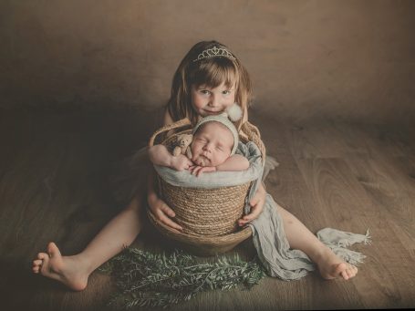 Niña abrazando a un bebé en una cesta sobre un fondo neutro.