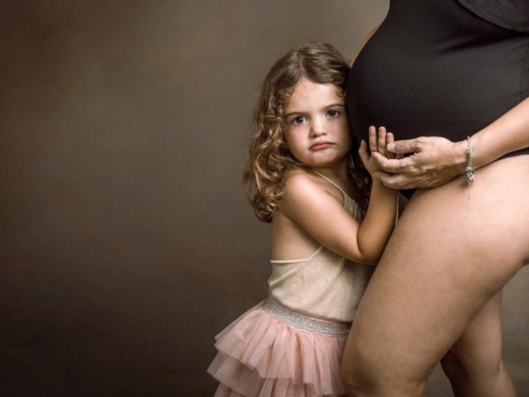 Niña abrazando la pierna de una mujer embarazada, ambos con expresiones serias.