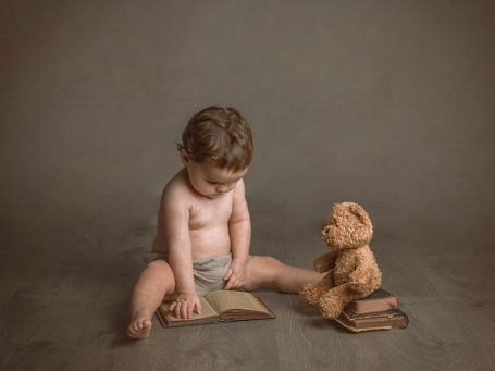 Niño pequeño sentado en el suelo, leyendo un libro con un osito de peluche a su lado.